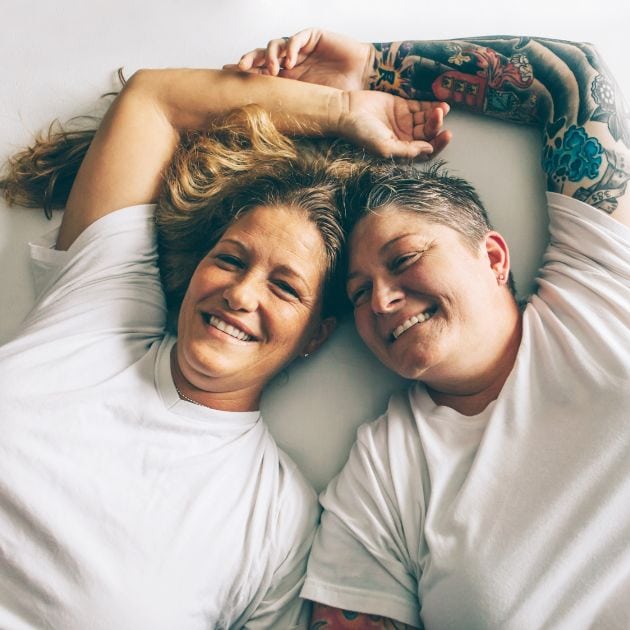 Two women are laying on a bed smiling. They are each wearing white tee-shirts and their hands are over their heads touching.