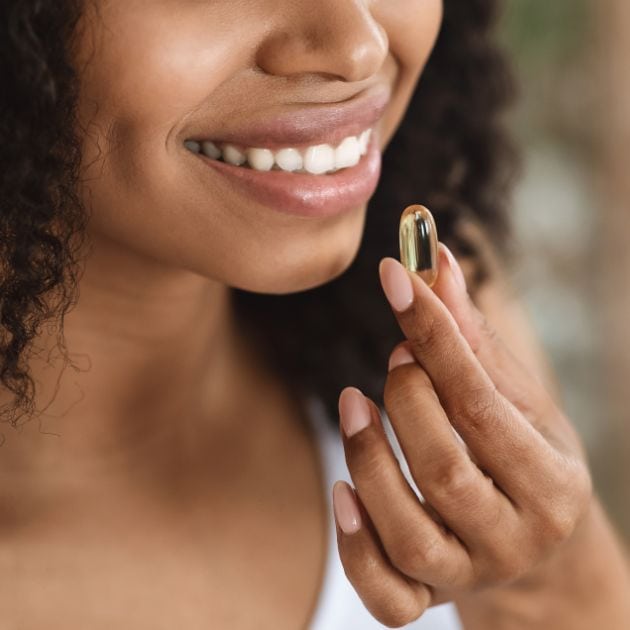 An African American woman is smiling and holding a vitamin up to her mouth.