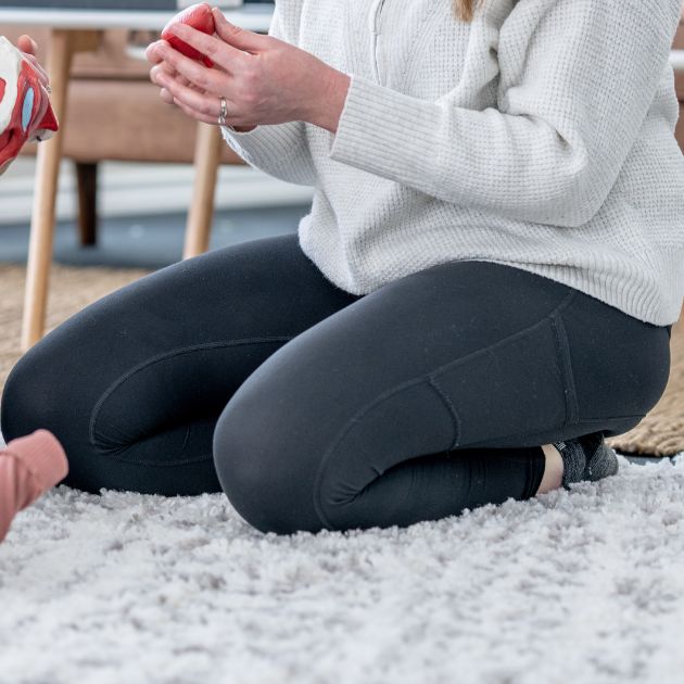 A woman wearing a light grey sweater and black leggings is sitting on her knees on a white shaggy rug.