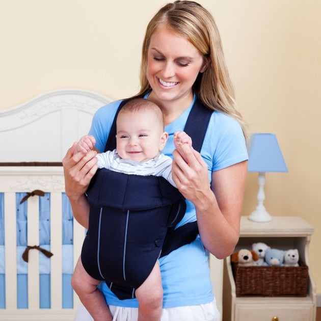 A woman wearing a bright blue teeshirt an white pants is wearing a baby in a harness. The baby is smiling.