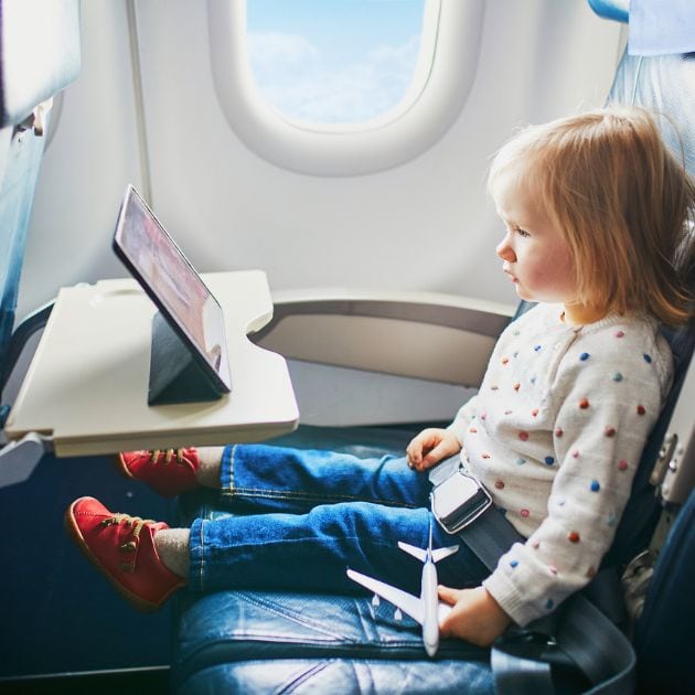 A little girl is sitting in a seat on an airplane. She is wearing red shoes, jeans, and a white sweater with pompoms that are red, green, blue, and pink on it. She is watching an IPad.