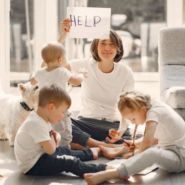 A woman is sitting on a hardwood floor. She has three kids all around her and a dog to the side. She is holding a sign that says "Help".