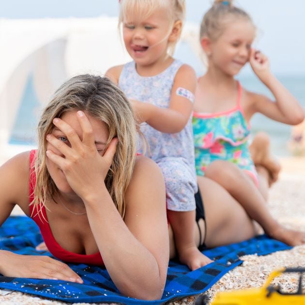 A woman in a black and red bathing suit is laying on a towel at the beach on her stomach. Two little girls are sitting on her back. The woman has her hand over her face.