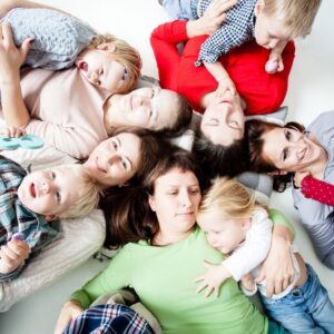 Five women are laying on the ground with their heads together in a circle. Each one has a child that is laying beside them.