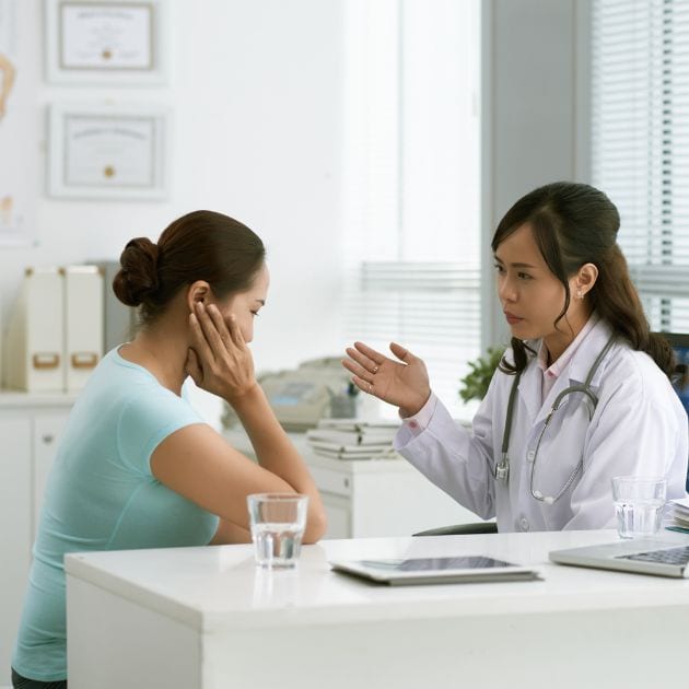 A woman wearing a seafoam green shirt is sitting at a doctor's desk. A female doctor is sitting across from her talking.