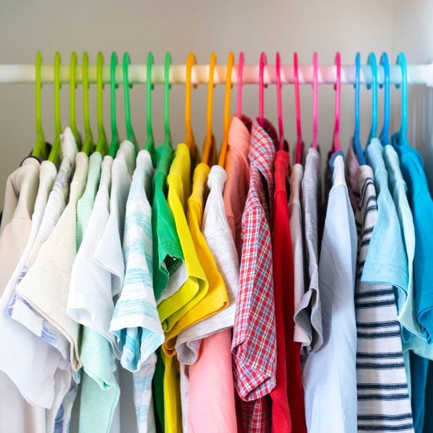 A kids closet holds shirts that are organized by colors, white, green, yellow, red, and blue. There are color coded clothes hangers as well.