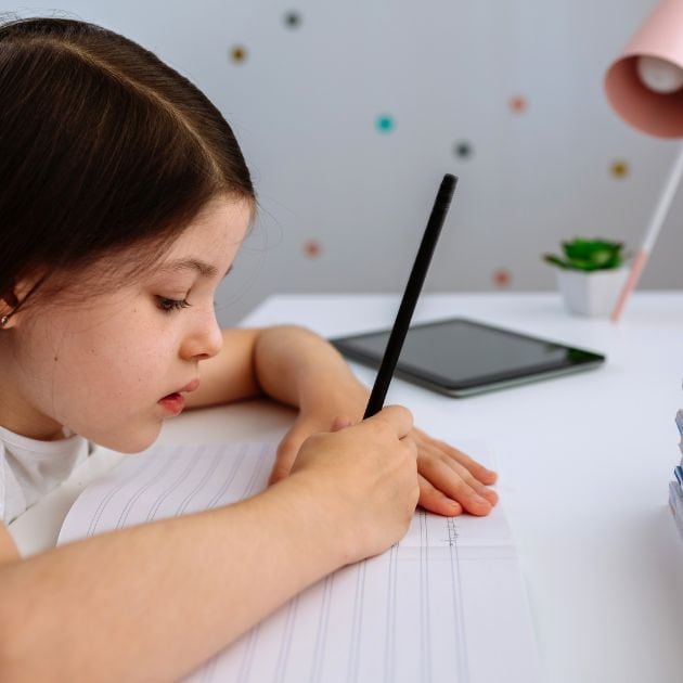 A girl with dark brown hair is sitting at a desk. There is an IPad and pink lamp to her left and she is holding a black pencil and writing on a piece of paper.