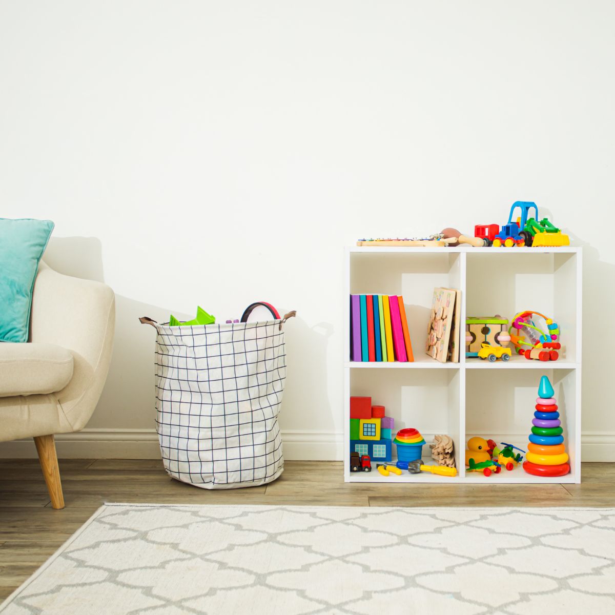 Square photo of a kid's room with a rug in the front of the picture. Behind the rug is part of a white chair with a blue pillow, a laundry bin with toys showing from the top, and a bookshelf being used as yor storage