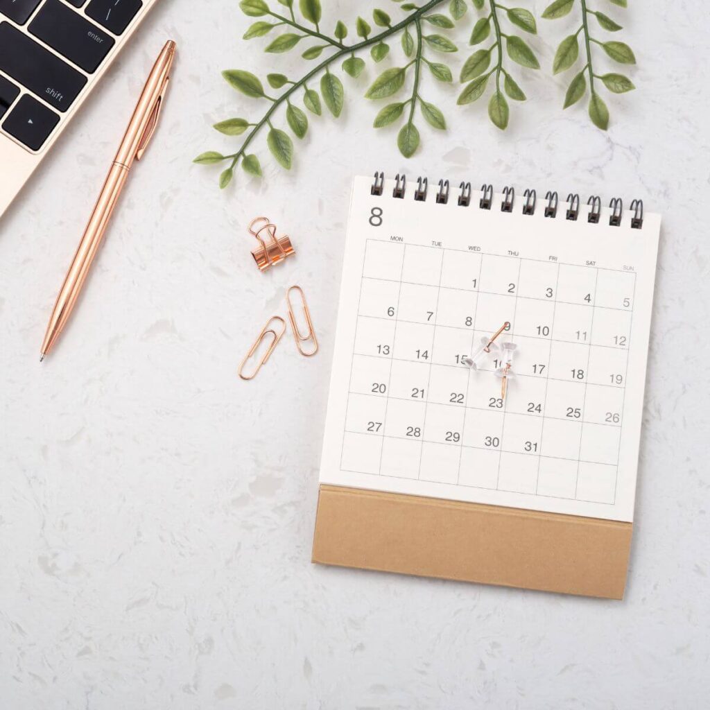 Flatlay photo on a white background of a calendar sitting on a desk with push pins on top. Above the calendar is a sprig of eucalyptus, and to the left are more push pins, a writing pen, and the corner of a laptop