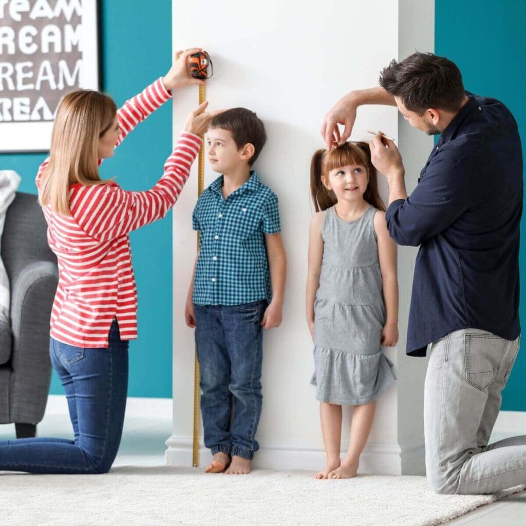 2 kids, a little boy and a little girl, stand with their backs against the wall while their parents use measuring tape to measure each of the kids' heights
