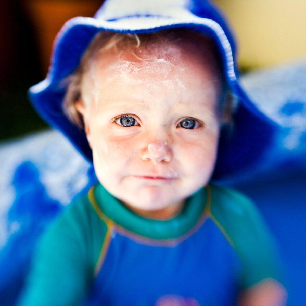 A boy is sitting down on a blue towel. He is wearing a blue and teal swim shirt and a dark blue hat.