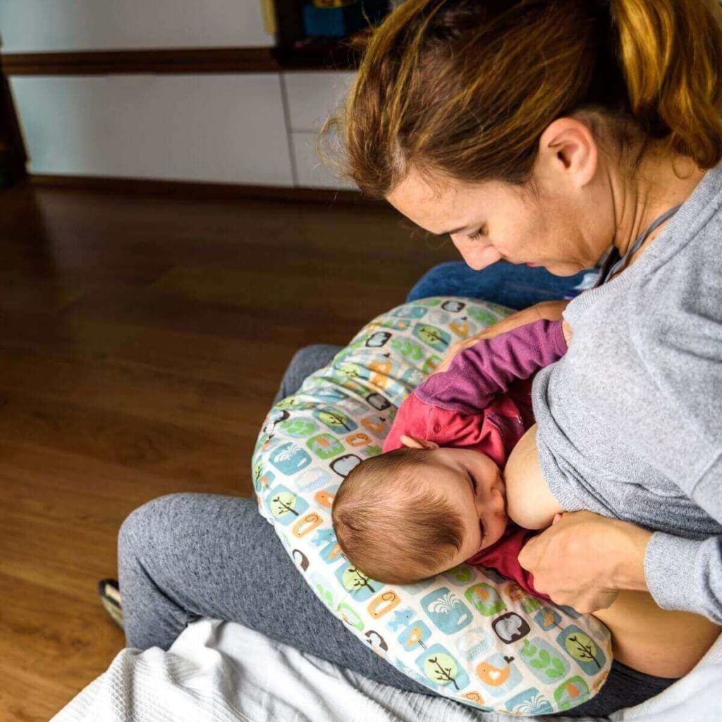 A woman in a light blue shirt is sitting on a couch. She has a nursing pillow around her waist and a little girl is laying on it while breasfeeding.