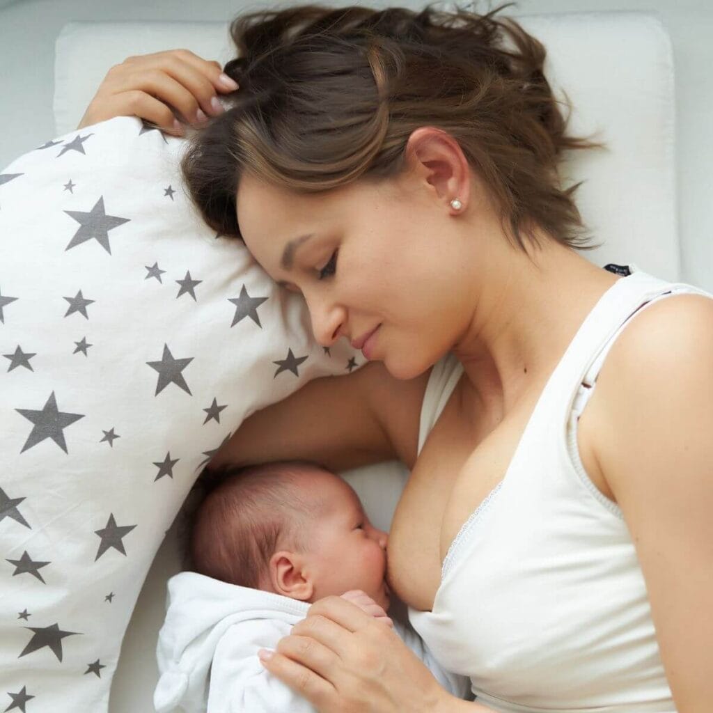 A woman in a white tank top is laying on her side on a bed breastfeeding her baby. Behind the baby is a white nursing pillow with dark grey stars.