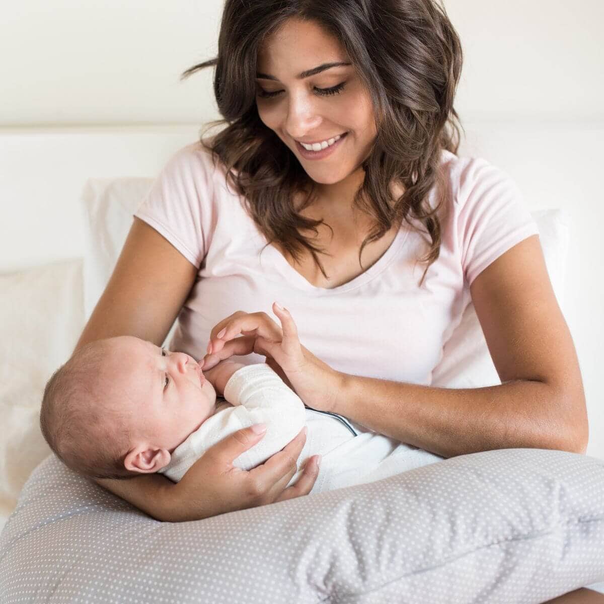A woman in a white shirt is sitting on a bed. She has a light grey nursing pillow with white polka dots in her lap while a baby lays on it.