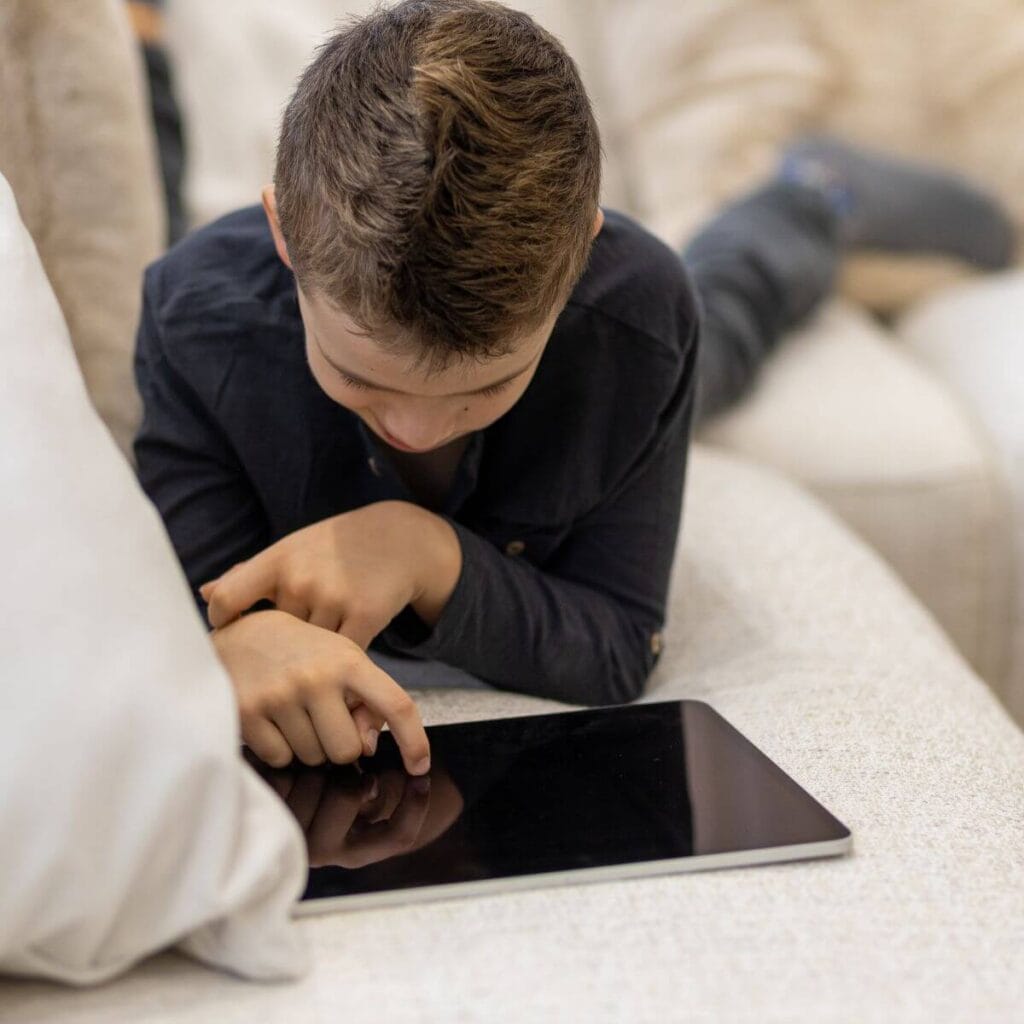 A boy wearing a black shirt is laying on his stomach on a white couch. He is playing on a tablet.