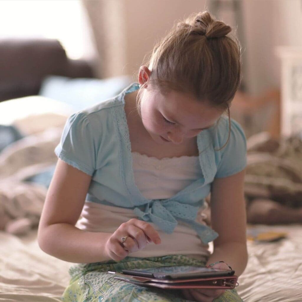 A girl with a white and blue shirt is sitting on the end of a bed looking down at a tablet.