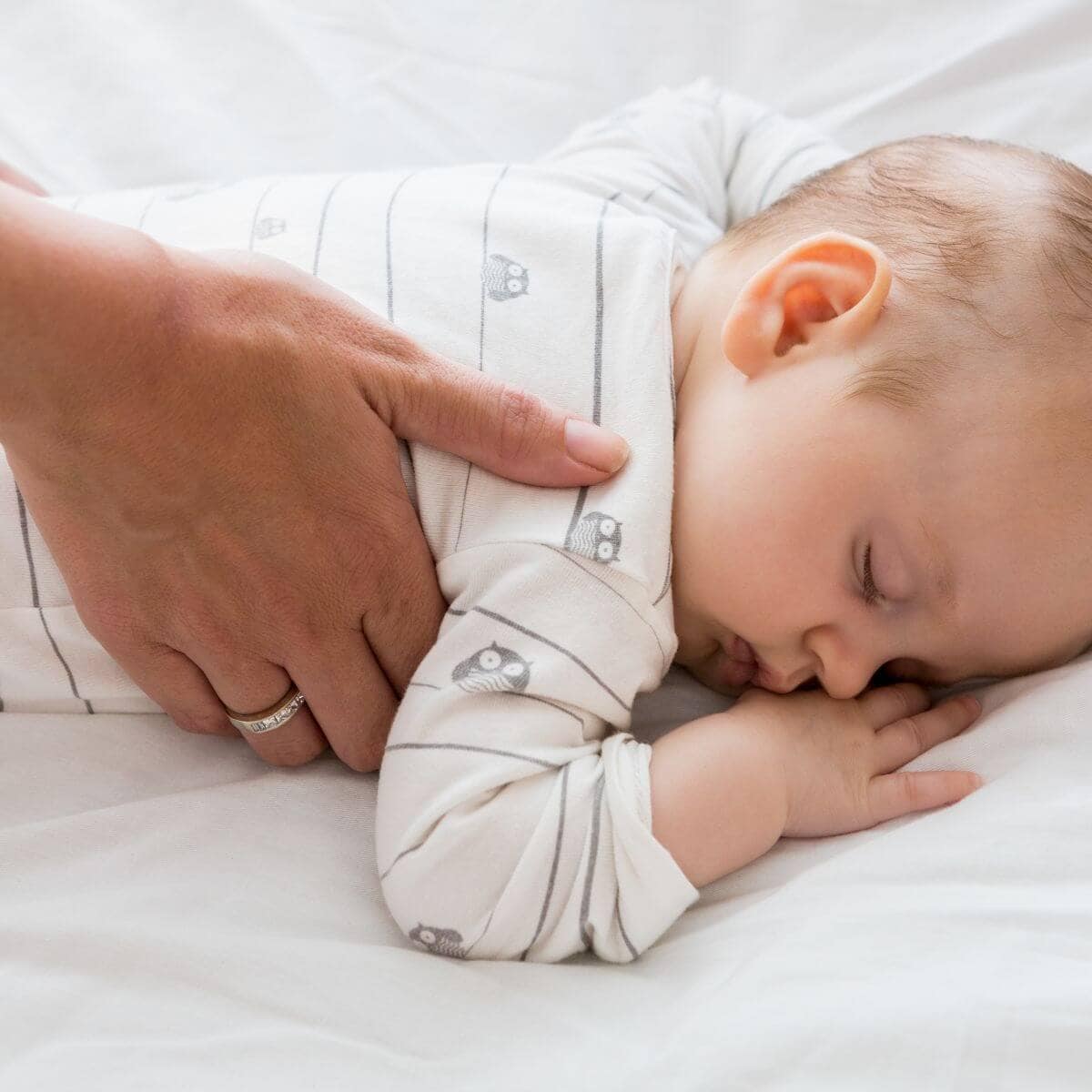 A baby boy is laying on his stomach sleeping on a white sheet. A man's hand is on the side of the baby.