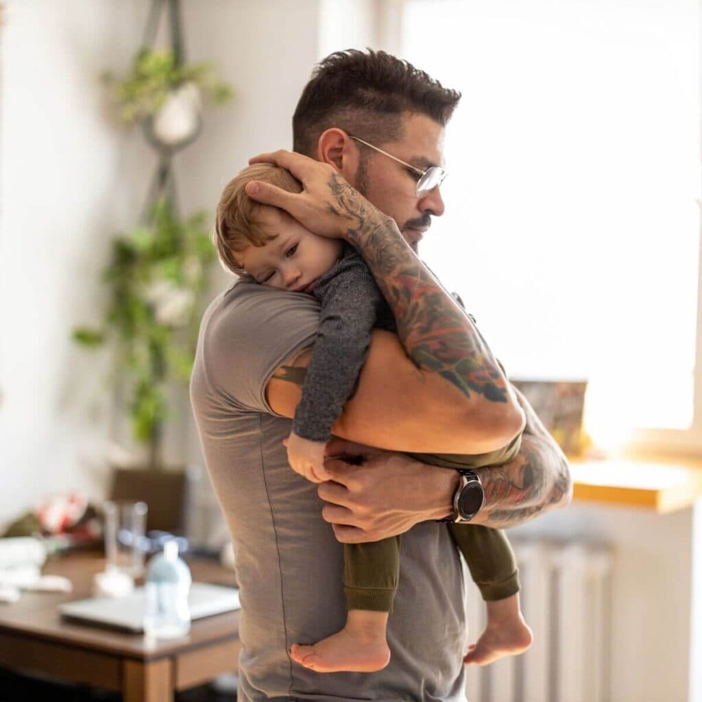 A man with tattoos on both arms is standing in the middle of a dining room holding a little boy who is almost asleep.