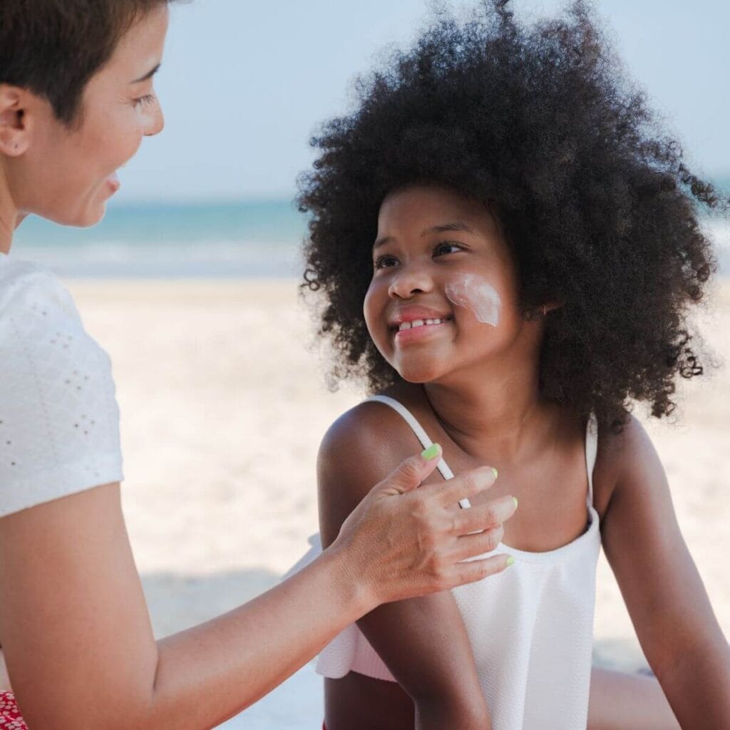 A girl with a white bathing suit on is sitting down on the sand with a woman. Both are smiling at eachother and the girl has sunscreen on her cheek.