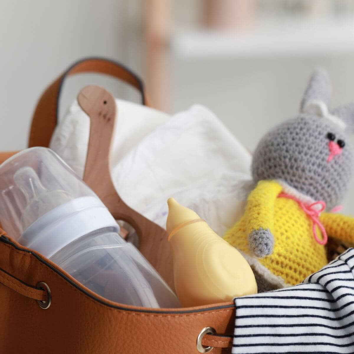 In a camel colored bag is diapers, a bottle, a nasal aspirator, a white and black striped outfit and a grey bunny with a yellow dress.