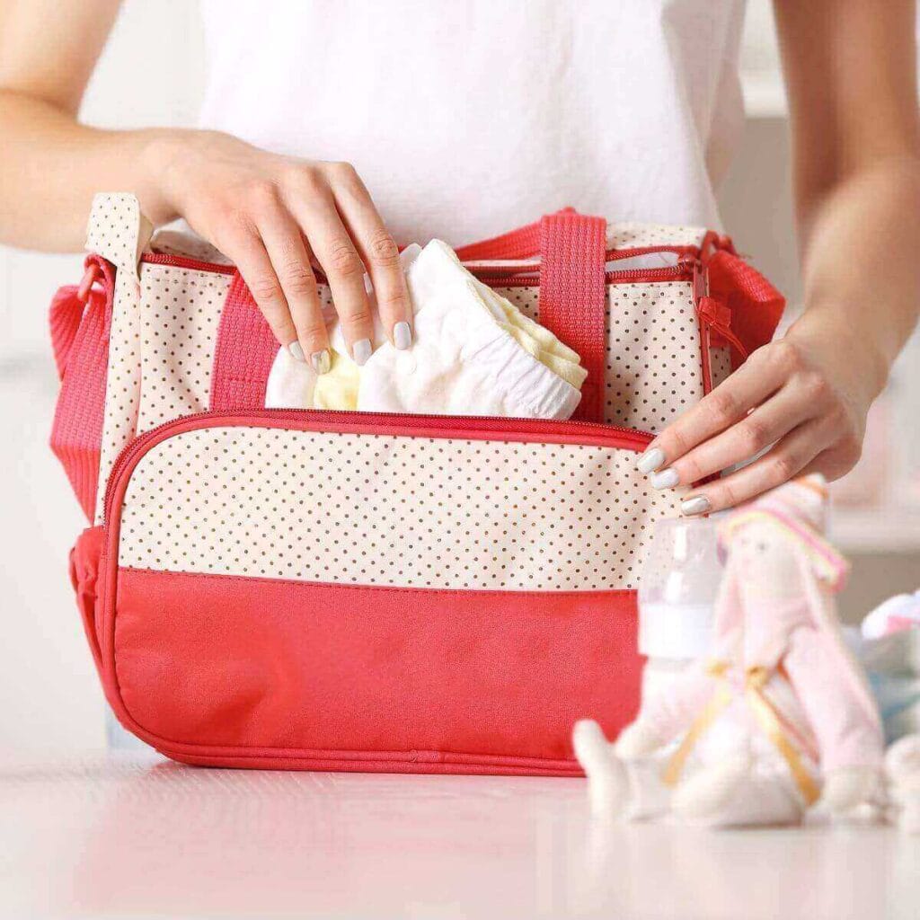 A woman is standing behind a table. On the table is a coral and white diaper bag. She is putting newborn pants in it.