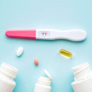 On a light blue table sits a pregnancy test and three white pill bottles. Over one is a white oval pill, over another is a pink oval pill and over the third is a yellow oval pill.