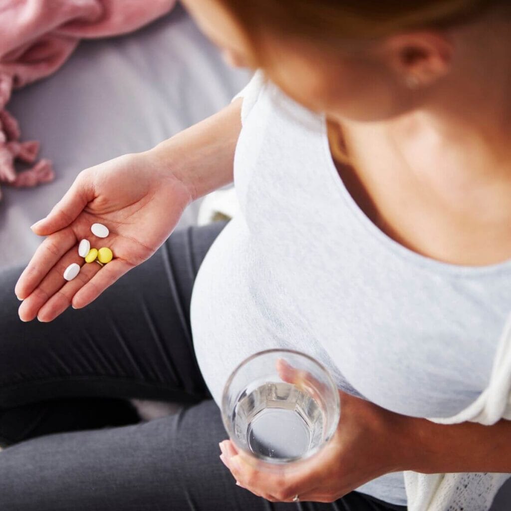 A pregnant woman is sitting on a light grey couch. She is holding 3 white and 2 yellow pills in her right hand and a glass of water in her left hand.