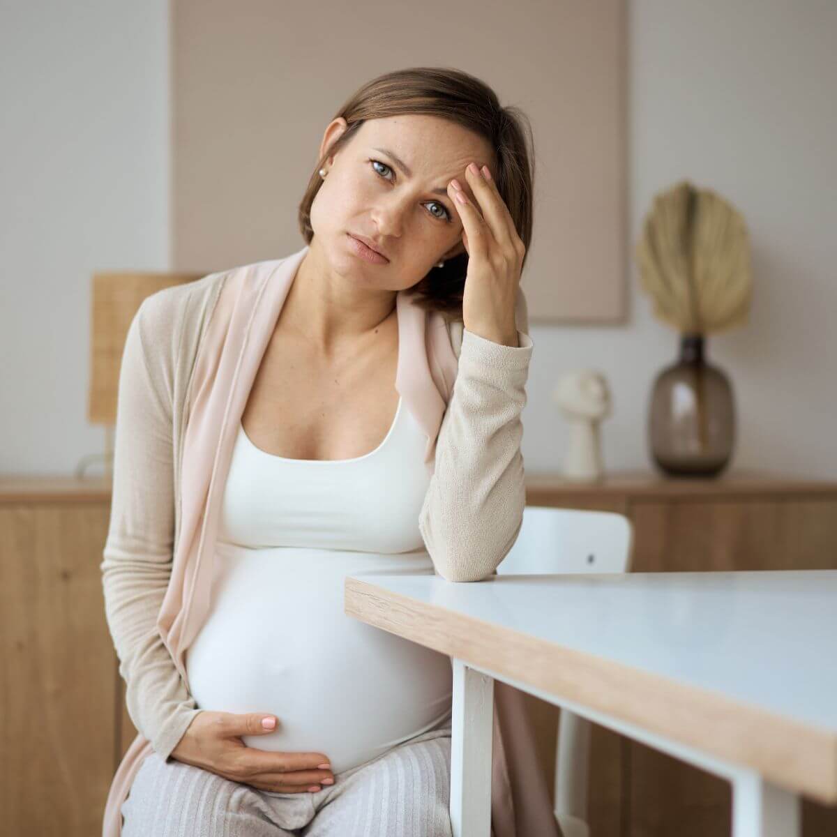A pregnant woman is sitting in a chair at a table. She has her right hand cupped under her stomach and her left hand on her forehead. She is staring into the camera.