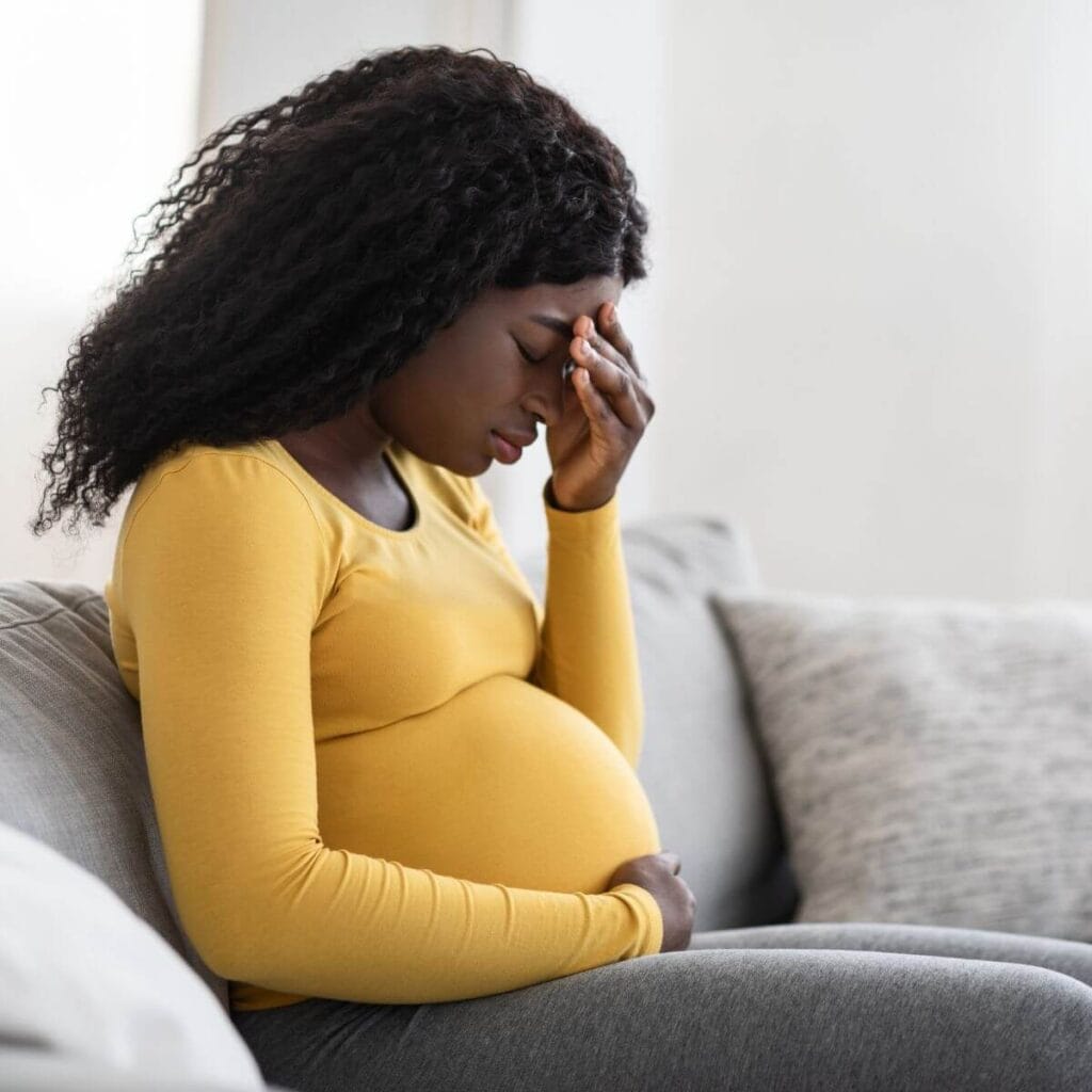 A pregnant woman is sitting on a light grey couch. She has her right hand cupping her stomach and her left hand is on her forehead. She has her eyes closed and looks sad.