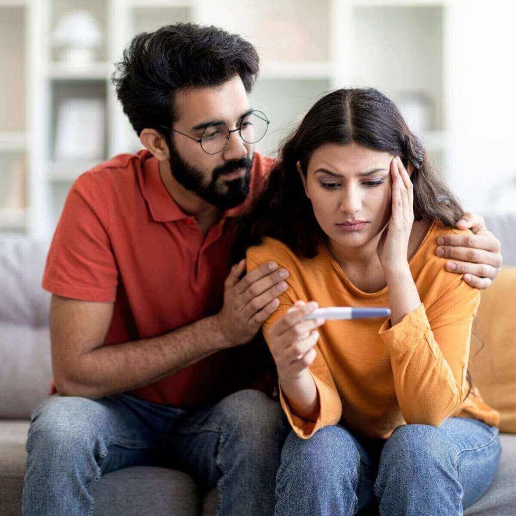 A man and woman are sitting on a light grey couch. They are both looking down at a pregnancy test the woman is holding and they look sad.