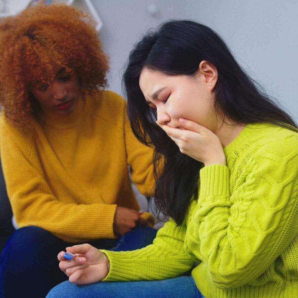 A woman in a yellow sweater is sitting with a woman in a green sweater. The woman in the green sweater is looking down at a pregnancy test with her hand over her mouth.