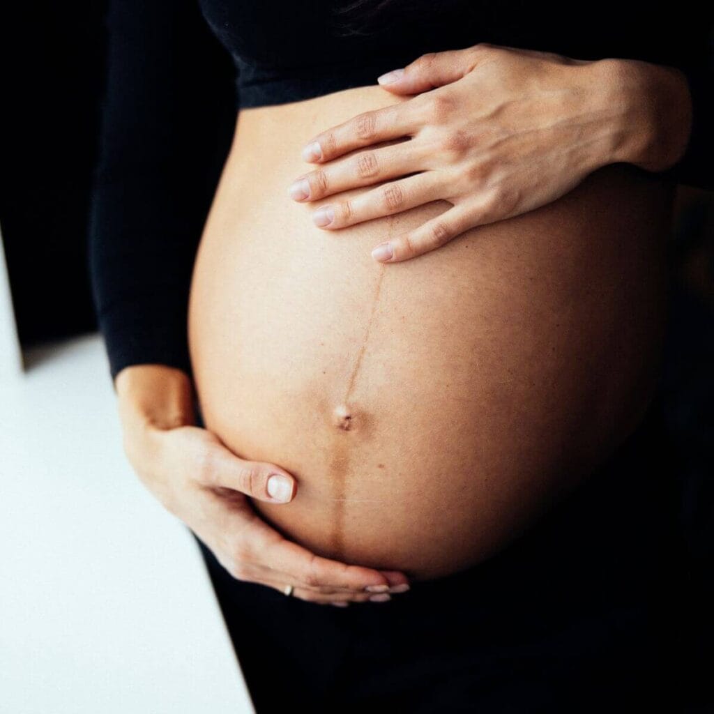 A close-up photo of a pregnant belly with a dark line called linea negra running vertically through the belly button. The woman in the photo is cupping her pregnant belly with her hands. 