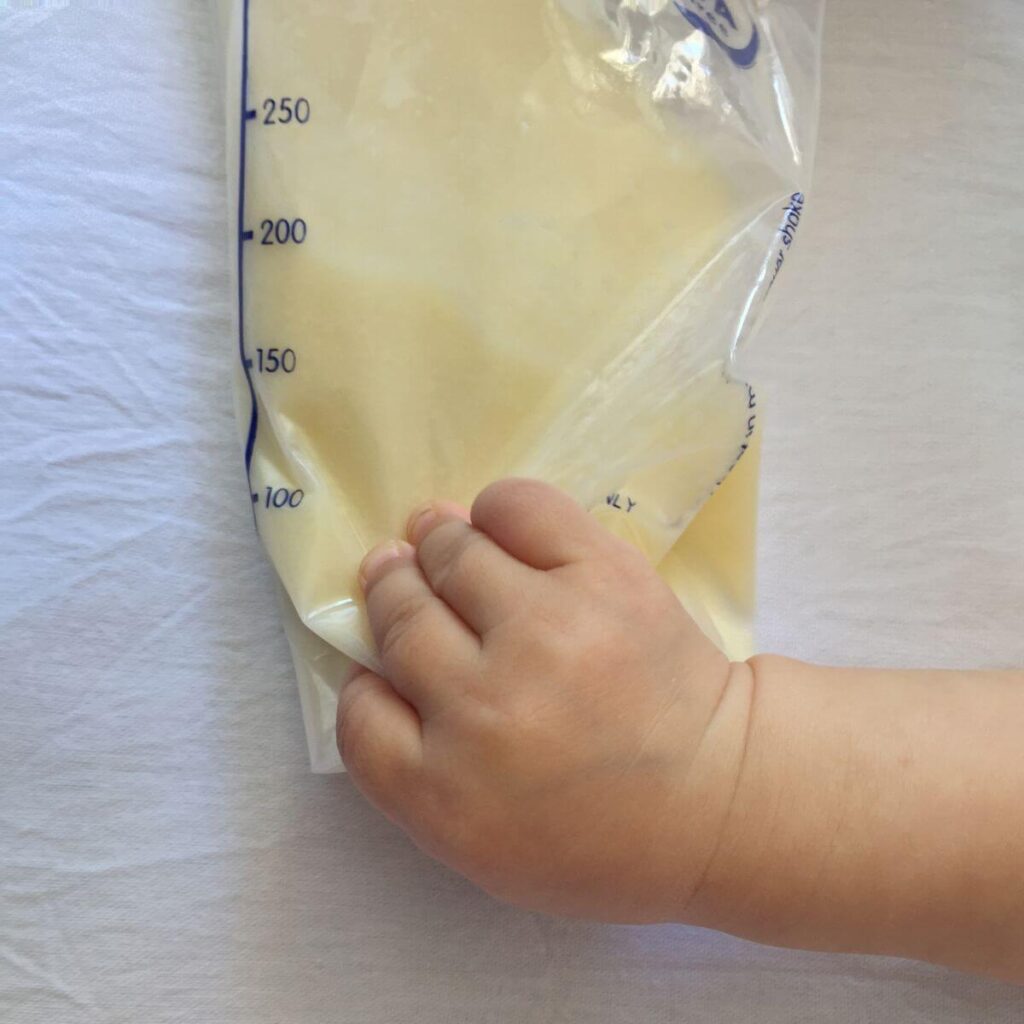 On a white sheet is a bag of breastmilk with a little ones hand holding it.