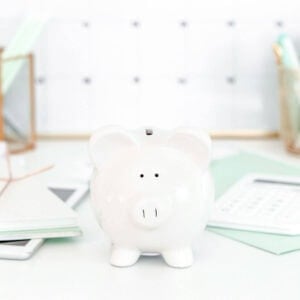 A square image focused on a white ceramic piggy bank in the center of the photo. The piggy bank sits on a white surface with notepads on both sides that are a seafoam green color. In the background sits a calendar.