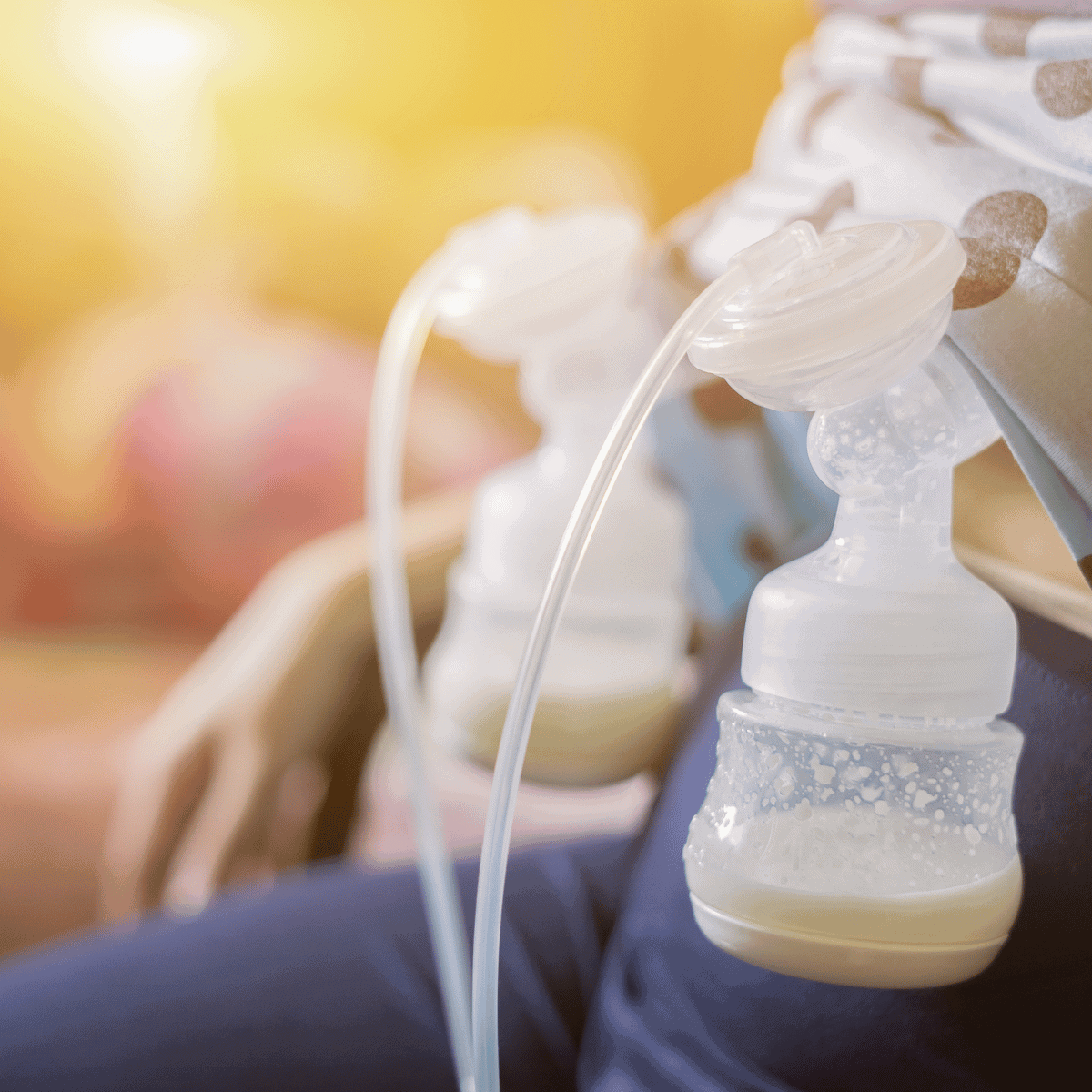A close-up image of a breast pump in use. The pump is attached to a person who is out of focus in the background. Milk can be seen collecting in the attached bottle, signifying the process of expressing breast milk. The environment is warm with a soft light that suggests a comfortable and private setting for pumping.