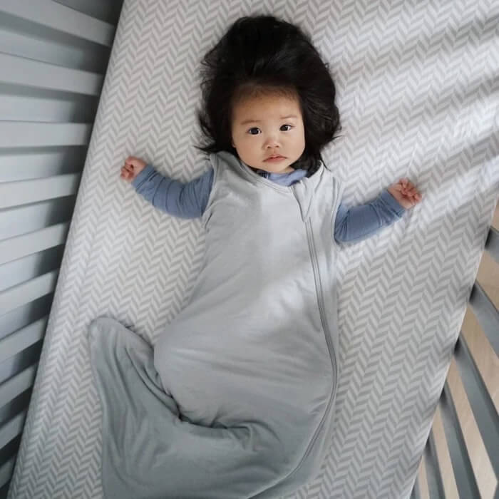 A toddler with a full head of hair lies on their back in a crib, looking up. They are wearing a two-tone Kyte Baby sleep sack, which is light gray with blue sleeves. The sleep sack is designed to keep them cozy and safe while sleeping. The crib mattress is covered with a chevron-patterned sheet, complementing the tranquil vibe of the scene.