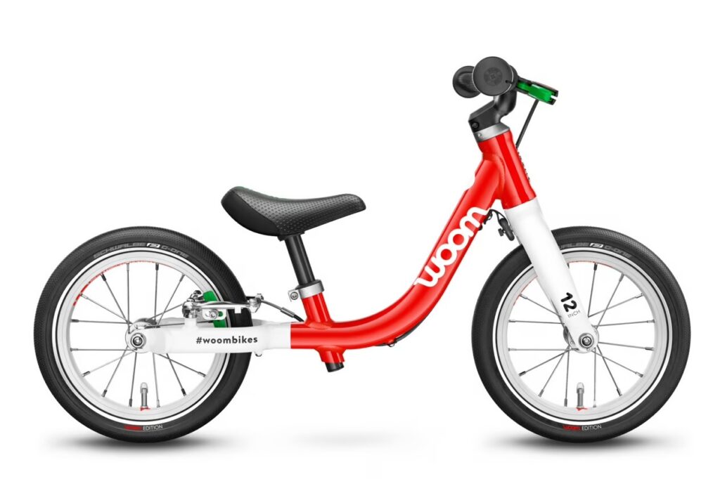 The image shows a children's balance bike from the brand "woom." The bike is predominantly red with a white front fork and features the "woom" logo prominently displayed on the frame. It has black tires with white rims and a silver-colored braking mechanism attached to the back wheel. The bike is designed without pedals, which is characteristic of a balance bike, intended to teach young children how to balance and steer before transitioning to a pedal bike.The bike also has a black, ergonomic saddle, and the handlebars are equipped with safety grips and a green brake lever, indicating the presence of a rear brake for added safety. The hashtag "#woombikes" is visible on the frame, suggesting that this image may be used for social media or promotional purposes. The overall design of the bike is modern and child-friendly, with an emphasis on safety and learning.