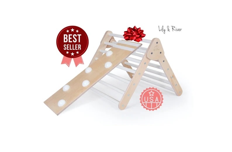 The image features a product called "Little Climber" by Lily & River, presented against a white background. It's a children's climbing frame made of wood, designed in a triangular A-frame shape with a runged ladder on one side and a smooth ramp with circular cut-outs on the other. The climbing frame has a natural wood finish, which gives it a clean and modern look suitable for indoor play spaces.Prominent on the image are two badges: one in red with white stars and text that reads "BEST SELLER," indicating that this item is a popular choice among consumers, and another badge resembling a seal with a red outline and the text "USA," likely signifying that the product is made in the USA. There's also a red ribbon bow on the top right, which suggests that this product could be a gift. The branding "Lily & River" is visible in a minimalist font, adding to the overall marketing of the product as high-quality and desirable.
