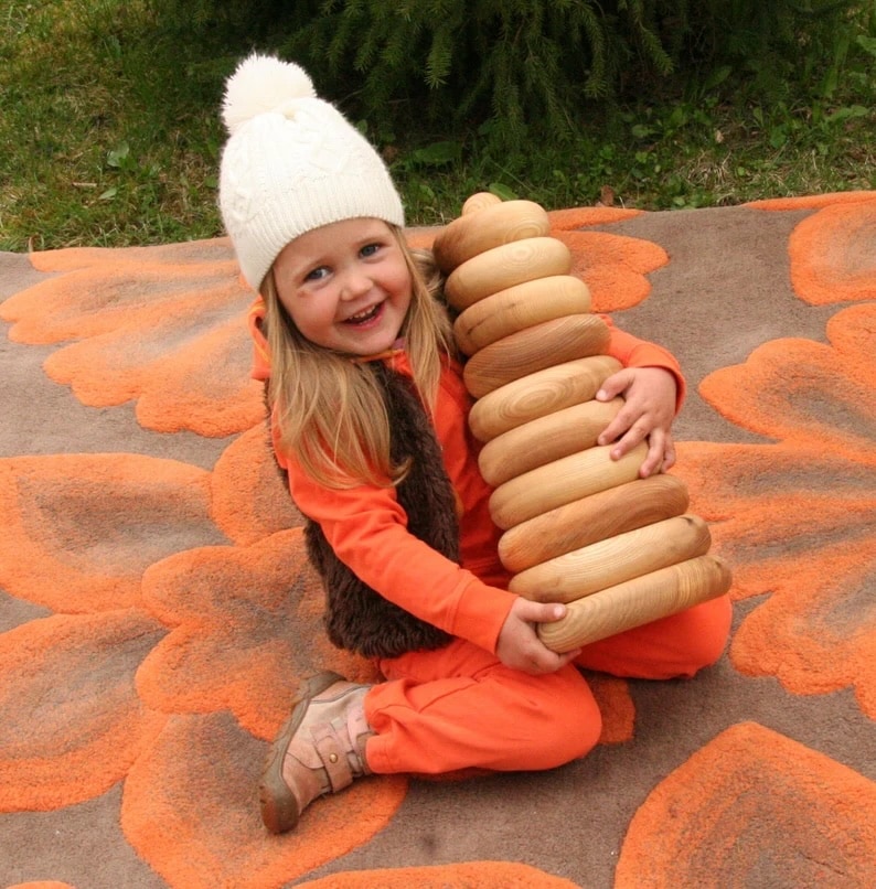 The image depicts a young child with a beaming smile, wearing a white knitted hat with a pompom on top and dressed in a bright orange outfit with a furry brown vest. The child is seated on an orange and brown floral patterned blanket on the grass, happily hugging a large handmade wooden stacking toy consisting of ten rings. The toy appears to be a Montessori or Waldorf-inspired educational toy, designed to encourage fine motor skill development and cognitive abilities in young children. The base of the toy is about 20 cm wide, indicating a substantial size for the stacking pyramid, which makes it an impressive and engaging toy for toddlers. The natural finish of the wood suggests a preference for eco-friendly and safe materials in children's toys.