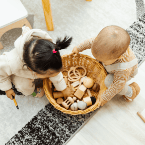 Two toddlers engaging with an assortment of wooden Montessori toys in a bright, cozy playroom. One child, with a playful pink hair tie, is kneeling and examining a toy closely, while the other, in a striped outfit, is reaching into the woven basket filled with blocks, rings, and spherical objects, encouraging tactile learning and exploration.