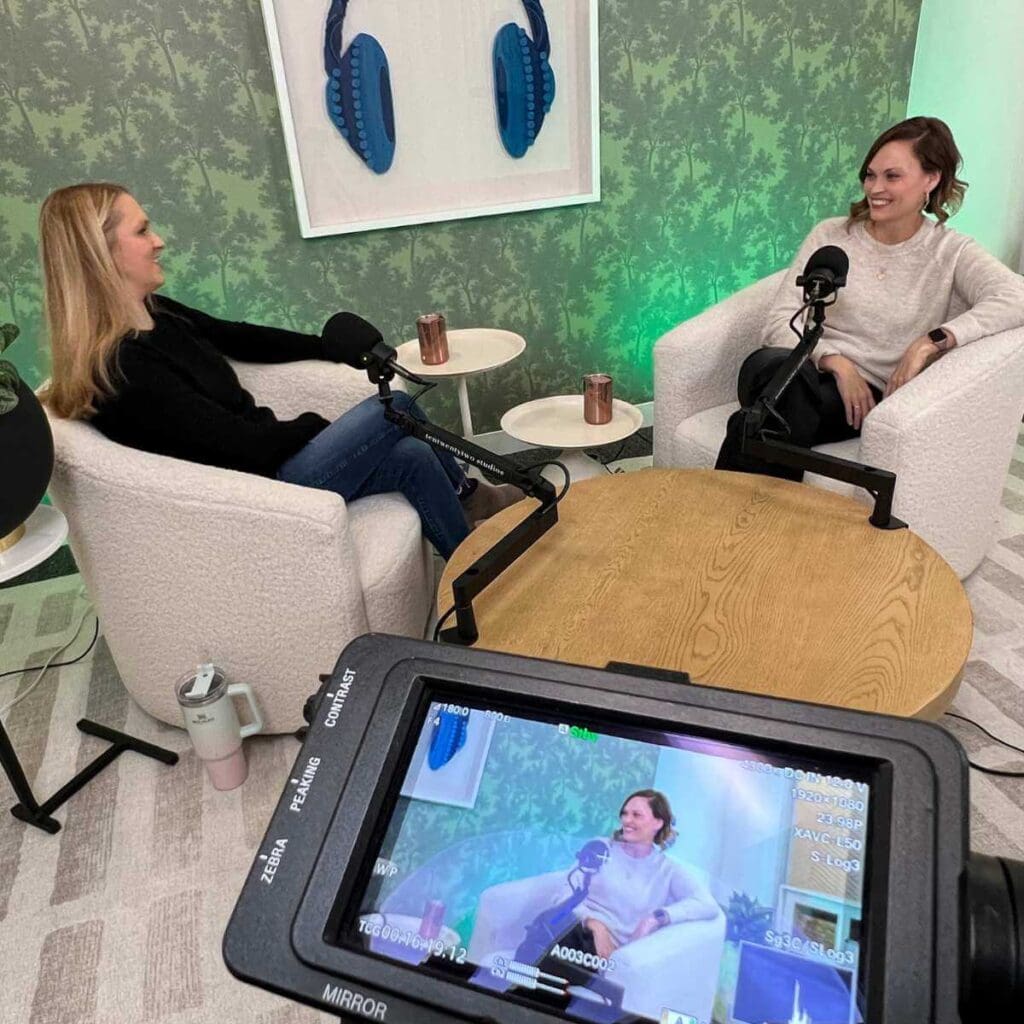 In a cozy podcasting setup for Undefining Motherhood, host Katy Huie Harrison and co-host Sarah Creel engage in conversation. They sit in comfortable white armchairs, separated by a wooden table, against a backdrop of green leafy wallpaper. A decorative blue headphone art piece hangs on the wall, adding a touch of podcasting theme to the decor.