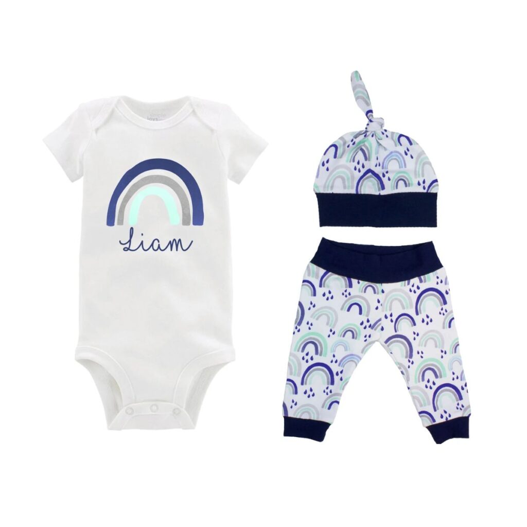 A white onesie with a blue-tone rainbow and the name "Liam" in blue script below the rainbow is on the left. To the right is a baby hat and a pair of baby pants with a blue rainbow motif.