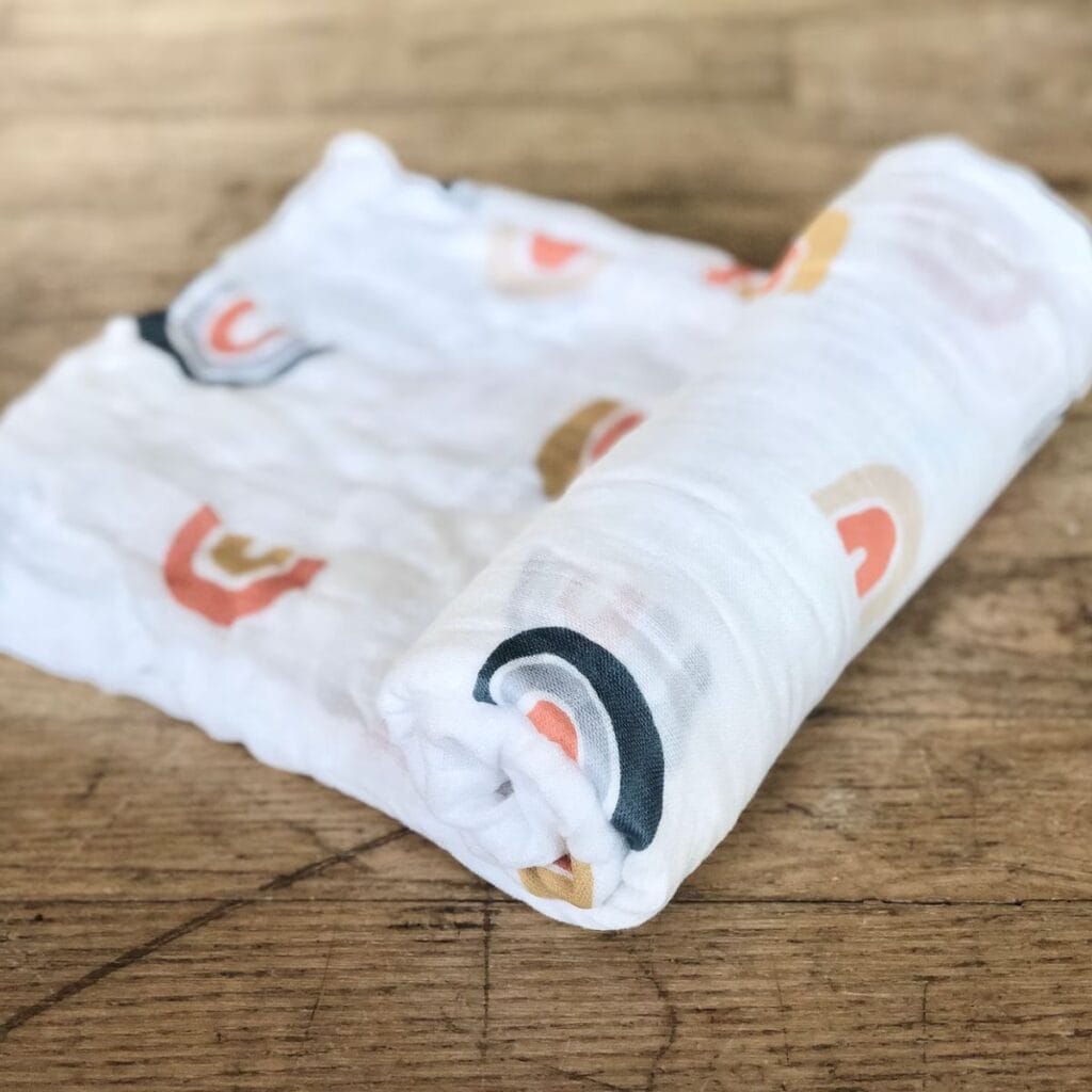 A muslin baby swaddle rolled up on a wooden surface. The swaddle is white but has earth toned rainbows decorating it.