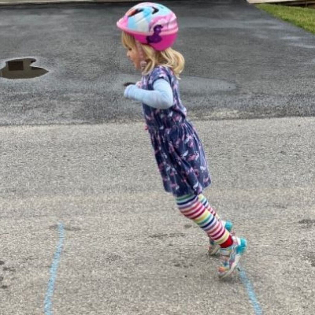Little girl jumping from one chalk line to next. She is wearing a pink helmet, rainbow striped leggings, a blue long sleeved t-shirt, and a navy short sleeve dress with flamingos on it.