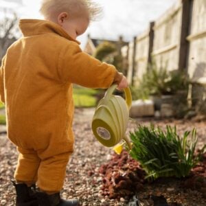 A toddler in a cozy mustard-colored onesie is watering plants with a lime green watering can. The child is standing to the side with their back partially turned to the camera, focusing on the task at hand. The sunlight illuminates the scene, highlighting the texture of the onesie and the child's light-colored hair. In the background, the garden pathway leads to a traditional building, suggesting a domestic outdoor setting.