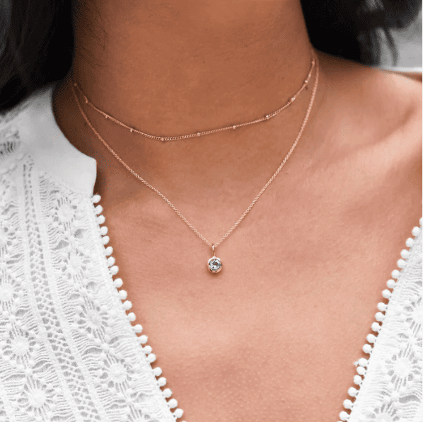 Picture of woman's neck in a lacy v-neck blouse. She is wearing a birthstone charm necklace from Tiny Tags and has another, shorter chain necklace layered with it.