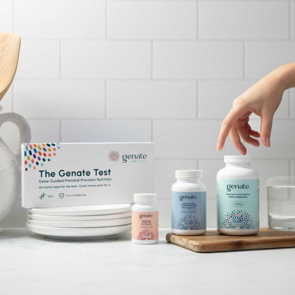 A counter displays The Genate Test kit for gene-guided prenatal precision nutrition, which includes a box labeled 'DNA' and 'CLIA-Certified Lab,' next to two supplement bottles, one labeled for essential prenatal nutrition and the other for dairy supplement. A person's hand is reaching for the dairy supplement bottle. Stacked white plates, a wooden cutting board, and two glasses of water complete the neat, health-conscious setting.