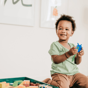 A joyful toddler with curly hair and a light green shirt smiles while holding a blue toy star. The child is seated next to a tray filled with colorful wooden blocks and shapes. The background is a clean, white room adorned with framed artwork, creating a bright and playful atmosphere.