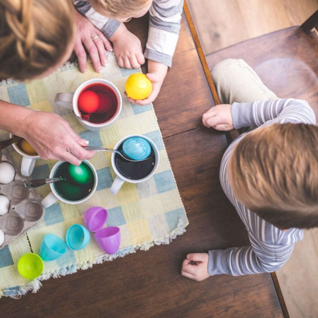 Two toddlers and a woman dyeing easter eggs in white mugs. One toddler is looking on from the side, while another holds a freshly dyed yellow egg in his hand.