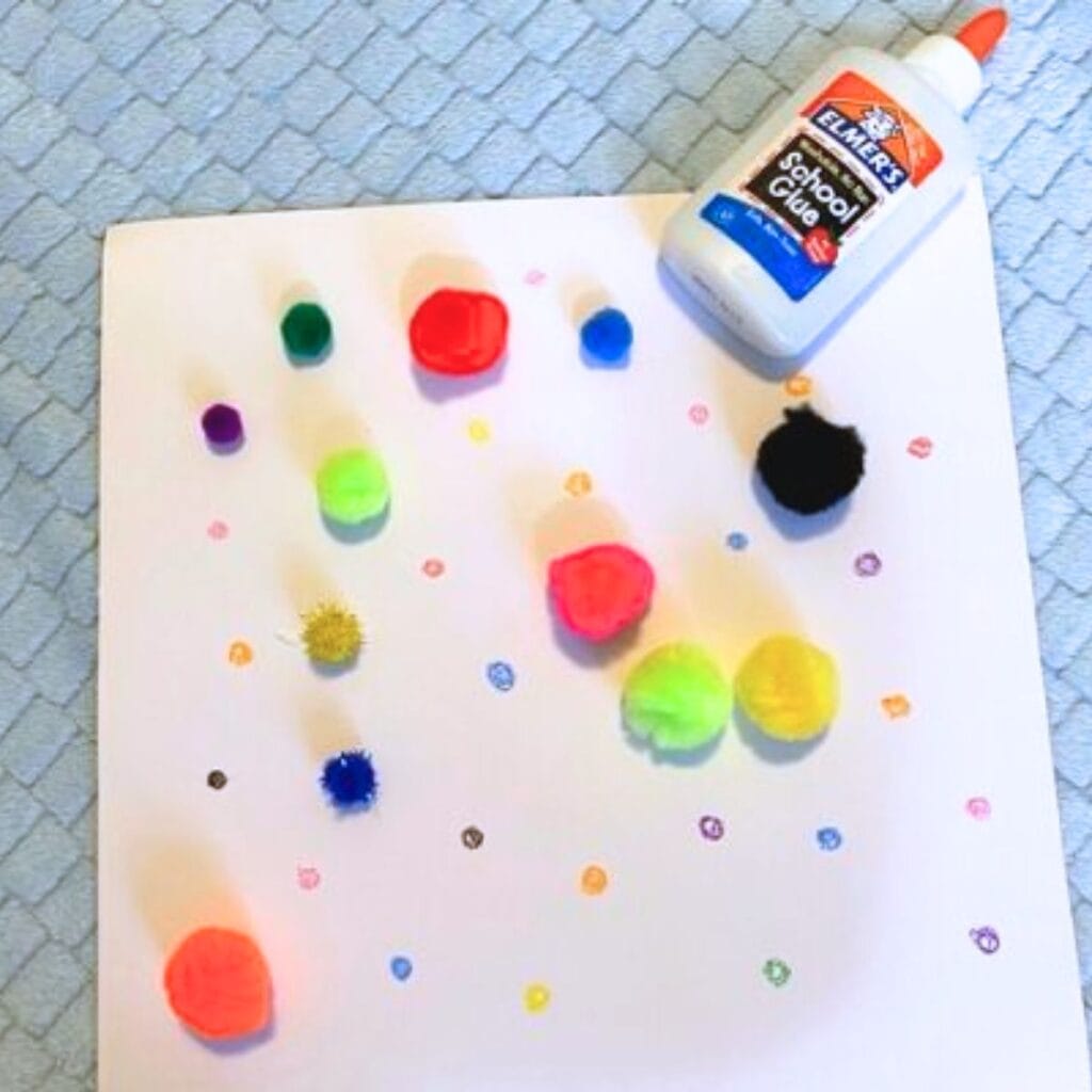 A white piece of computer paper sitting on a powder blue blanket background. The paper is covered in small dots of different colors, drawn on by colored pencil. There are several pom poms of different sizes and colors sitting on top of the paper, as well as a bottle of glue to the side.